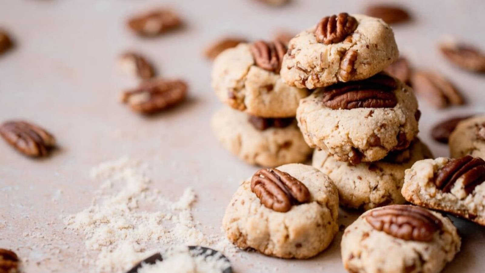 A stack of pecan cookies sits on a light surface, surrounded by whole pecans and cookie crumbs.