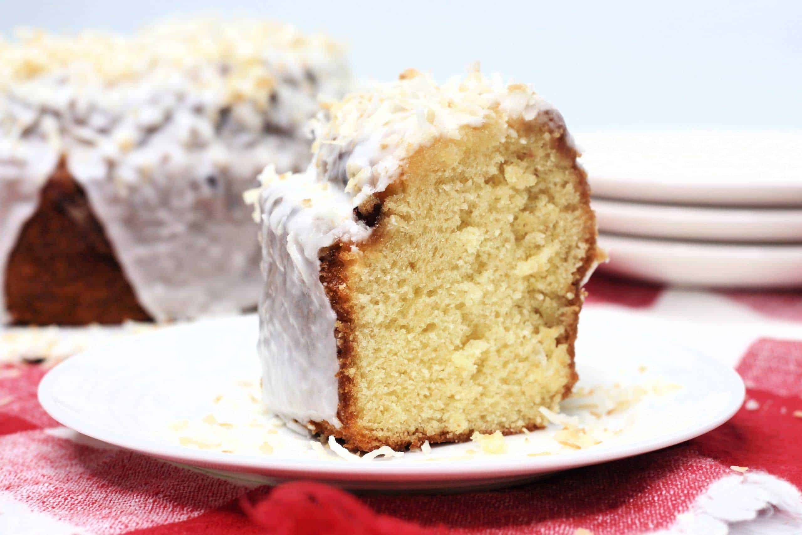 A slice of glazed bundt cake with shredded coconut on top is served on a white plate, with the rest of the cake and stacked plates in the background.