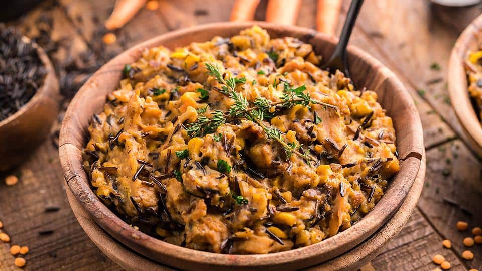 A wooden bowl filled with a creamy mixture of wild rice, corn, and vegetables, garnished with herbs, on a rustic wooden table.