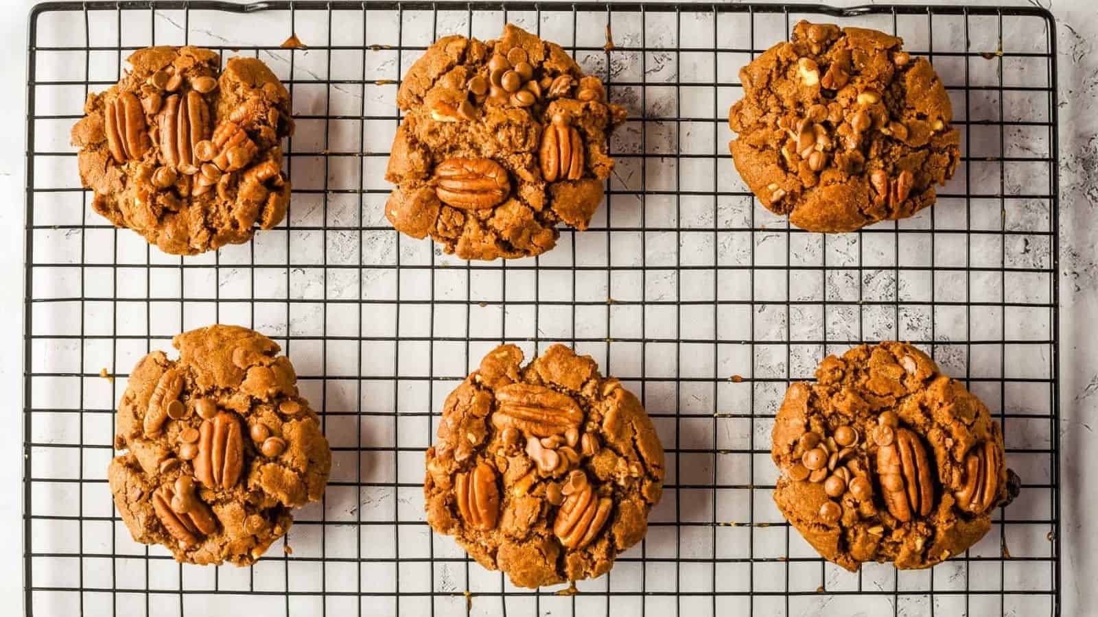 Six pecan and chocolate chip cookies cooling on a black wire rack, viewed from above on a white surface.