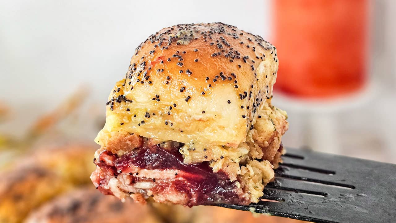 A close-up of a sandwich slider filled with meat, cranberry sauce, and melted cheese, topped with poppy seeds, held up on a spatula.