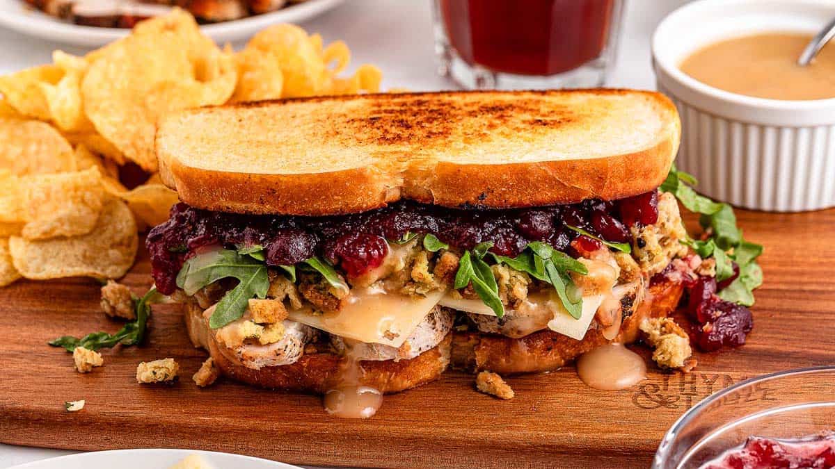 A turkey sandwich on toasted bread with stuffing, cranberry sauce, gravy, and leafy greens, served on a wooden board with potato chips and a cup of sauce in the background.