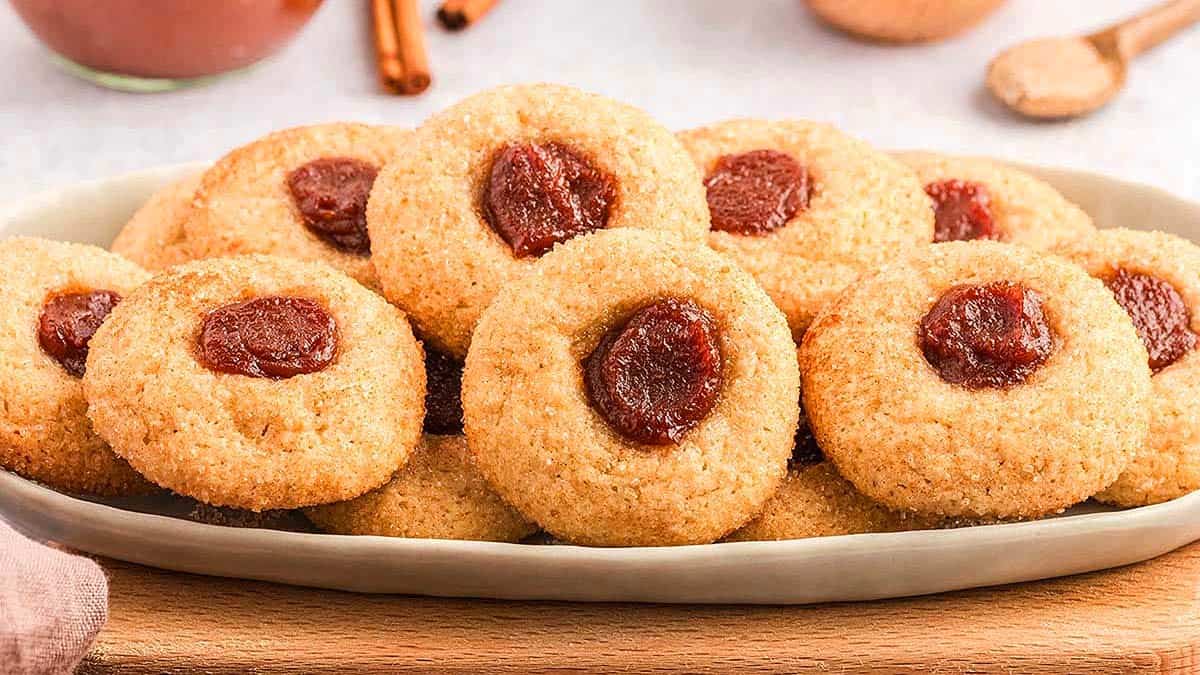 A plate of round cookies with a red jam center, arranged in a pile on a ceramic dish on a wooden surface.
