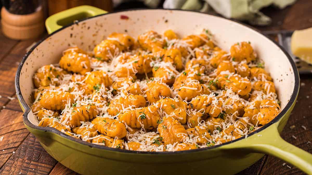 A green skillet filled with cooked gnocchi, topped with grated cheese and herbs, sitting on a wooden surface.