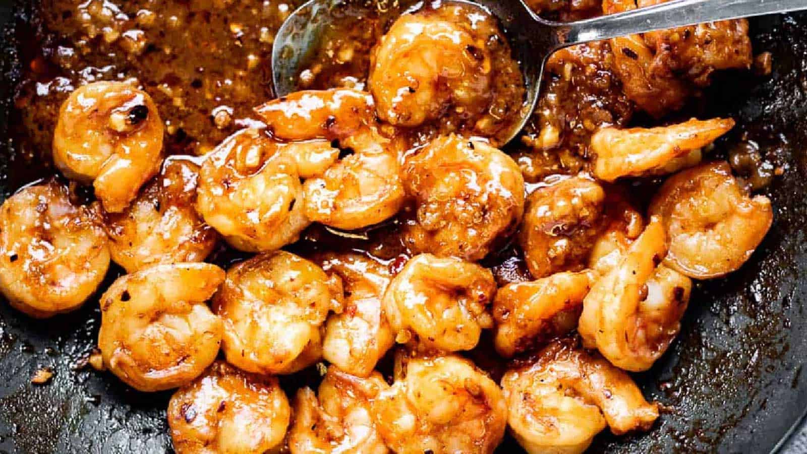 Cooked shrimp in a dark, glossy garlic sauce with visible seasonings, being stirred with a metal spoon in a skillet.