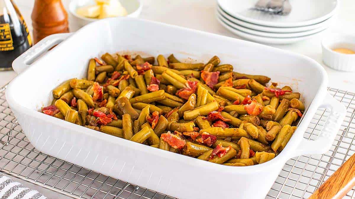 A white baking dish filled with cooked green beans and pieces of bacon sits on a cooling rack, with plates and utensils in the background.