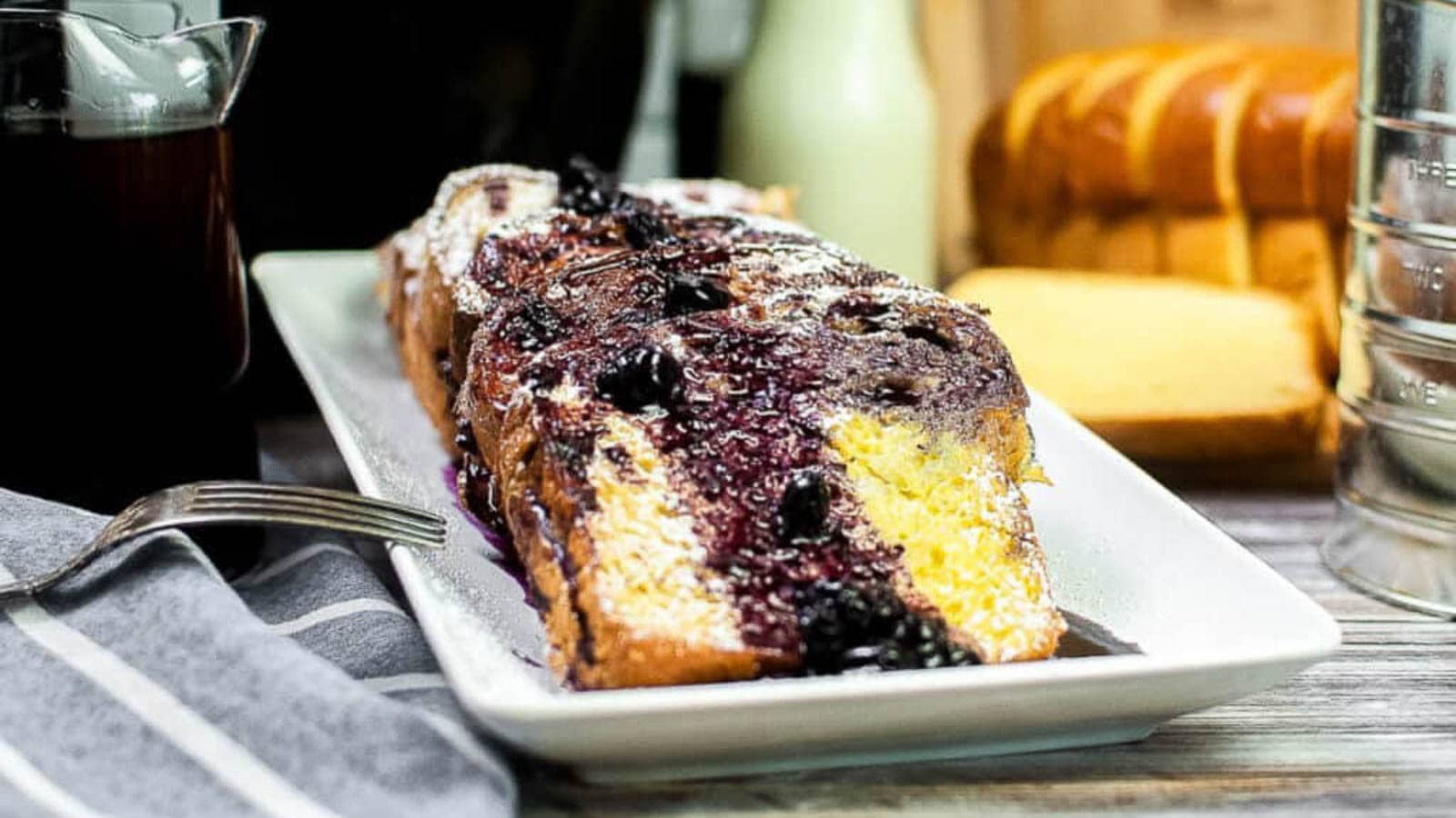 A rectangular loaf of blueberry bread dusted with powdered sugar sits on a white plate, with a sliced loaf and a pitcher in the background.