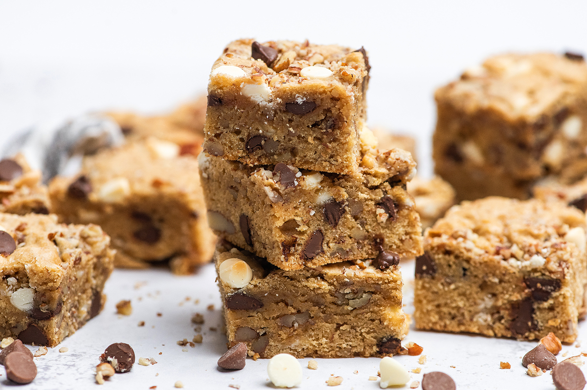 A stack of chocolate chip blondies with white and milk chocolate chips, surrounded by more blondies and scattered chocolate chips on a white surface.