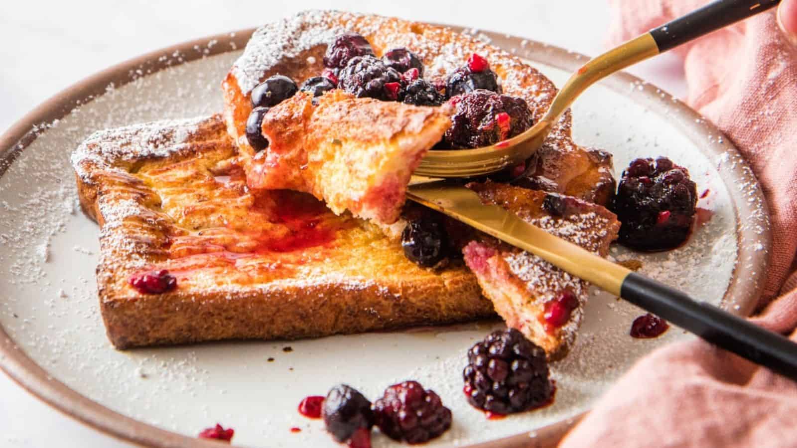 A plate of French toast topped with powdered sugar and mixed berries, being cut with a fork and knife.