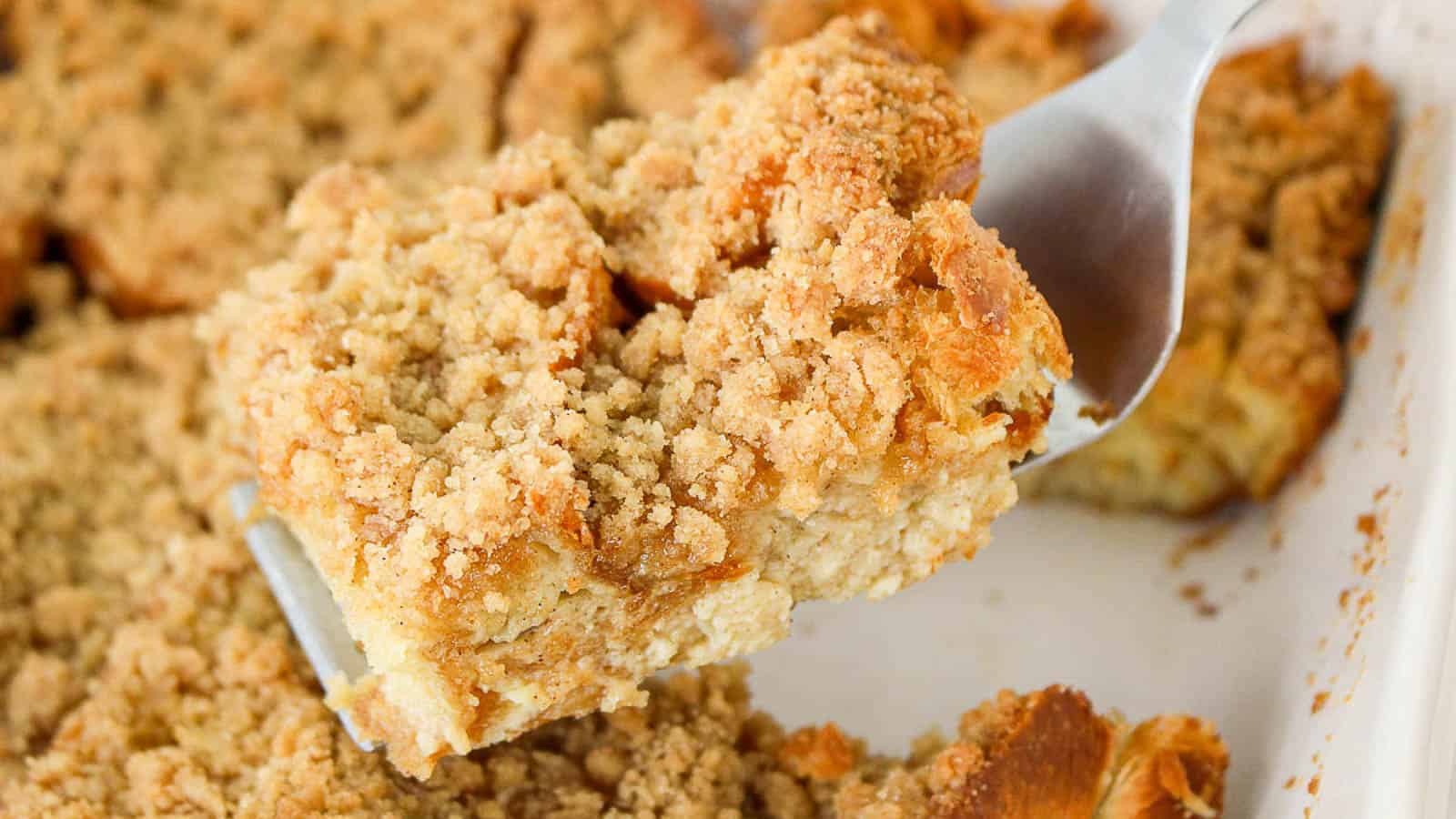 A metal spatula lifts a crumbly slice of baked dessert with a golden-brown streusel topping from a white baking dish.