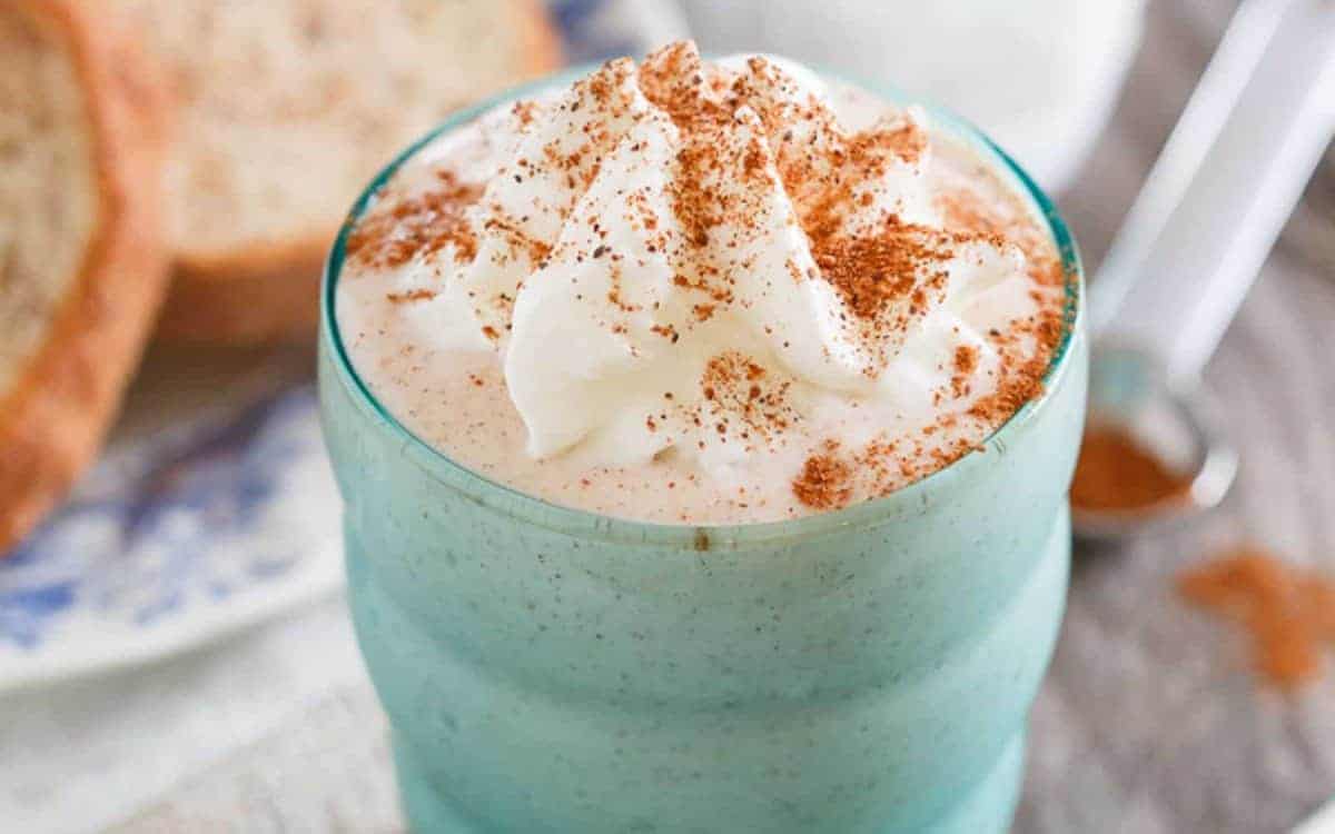 A glass filled with a creamy beverage topped with whipped cream and sprinkled with cinnamon, with bread and a spoon of cinnamon powder in the background.