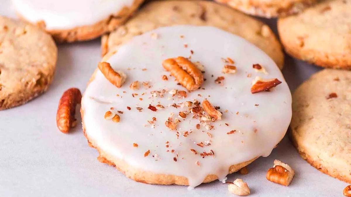 A close-up of cookies topped with white icing and sprinkled with chopped pecans, arranged on a light surface.