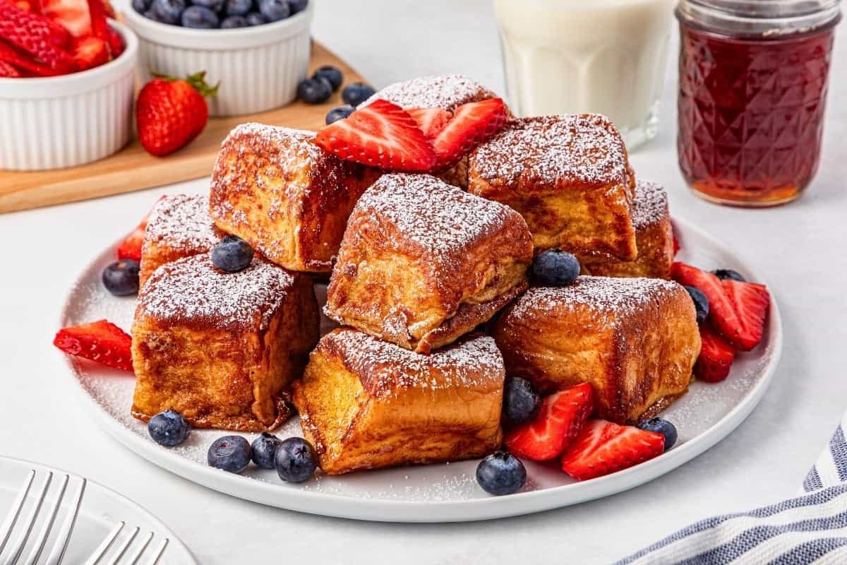 A plate of thick French toast cubes topped with powdered sugar, surrounded by sliced strawberries and blueberries. Milk and jam jars are in the background.