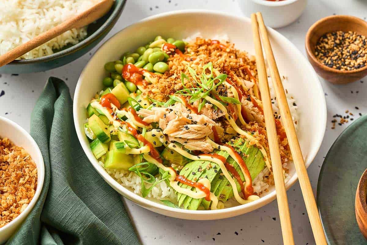 A poke bowl with white rice, shredded fish, sliced avocado, cucumber, edamame, crispy toppings, sauces, and green onions, served with chopsticks.