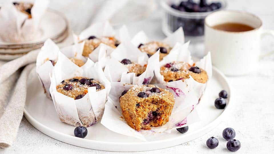 A plate of blueberry muffins in white paper liners sits next to a mug of tea, a bowl of blueberries, and stacked plates on a light table.