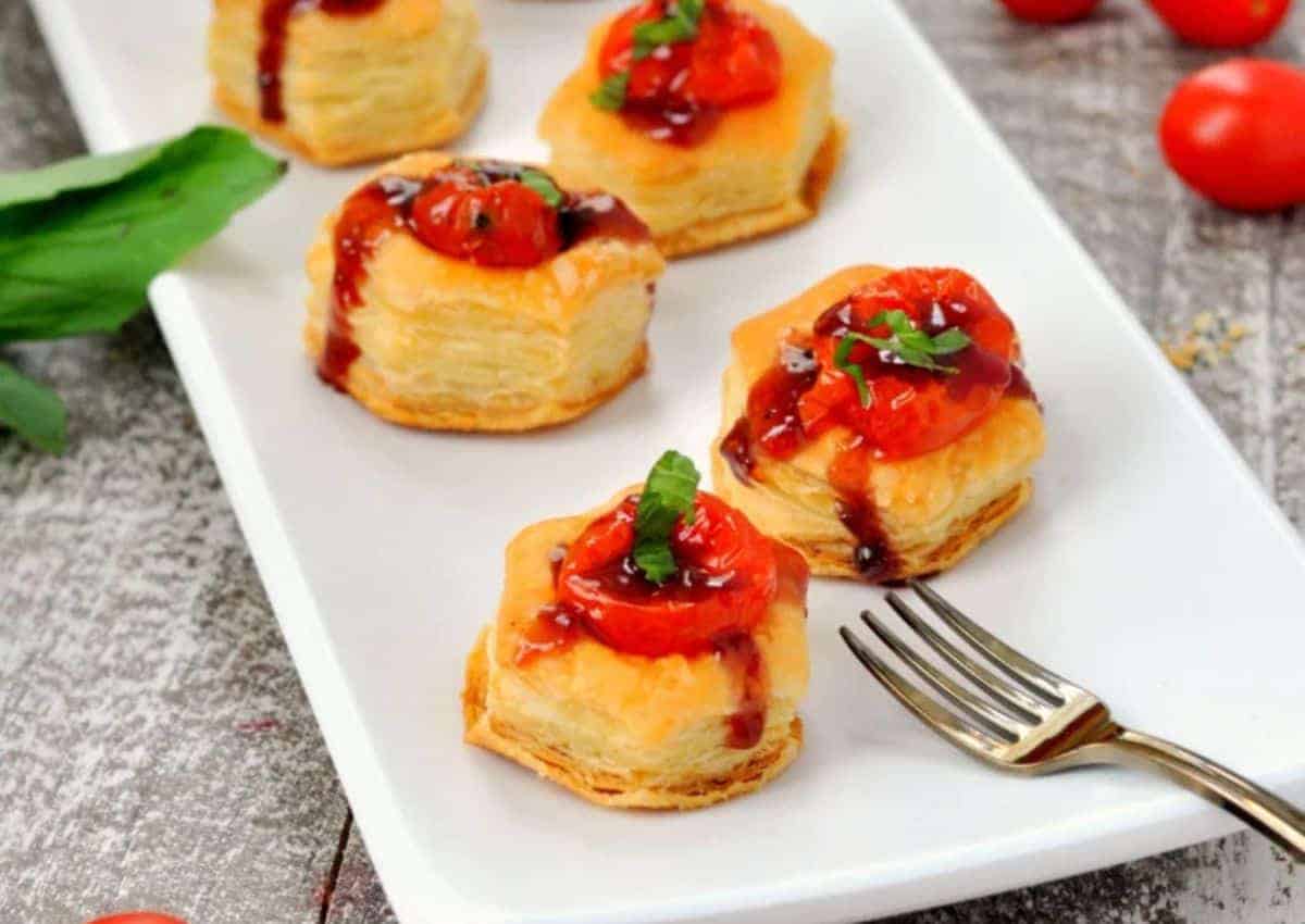 A white plate with puff pastry bites topped with roasted cherry tomatoes, balsamic glaze, and basil, next to a small fork and scattered cherry tomatoes.