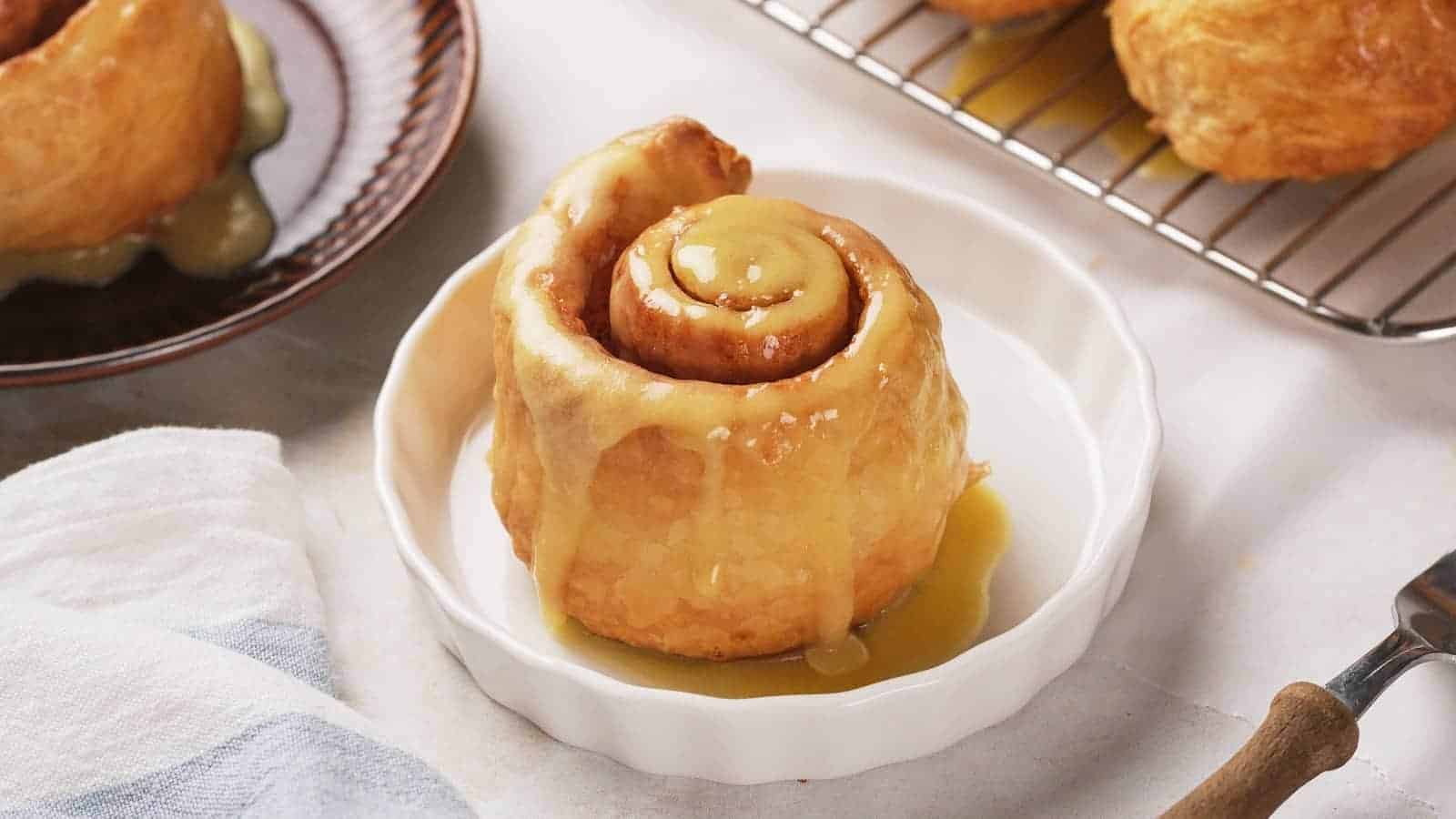 A cinnamon roll with glaze sits on a white dish, with more rolls on a cooling rack and a plate in the background.