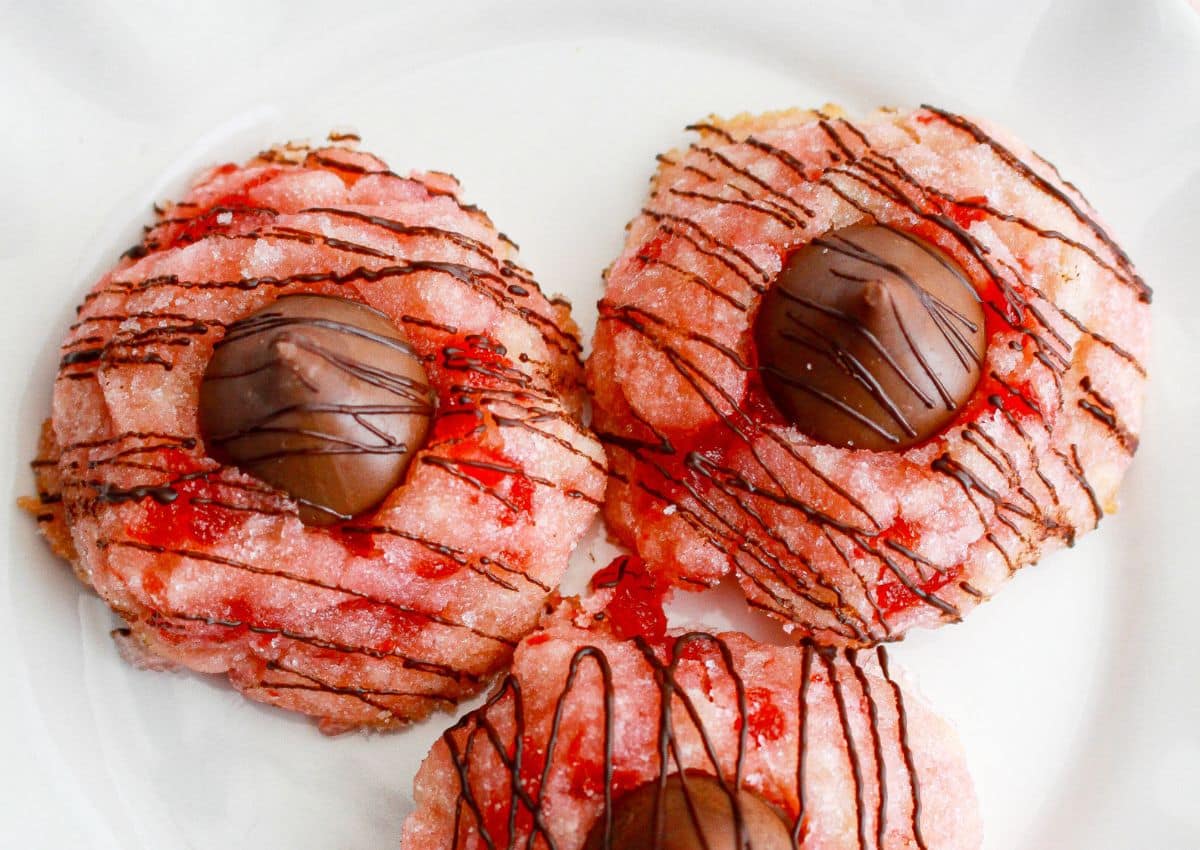 Three sugar-coated cookies with red bits, each topped with a chocolate candy and drizzled with chocolate, on a white plate.