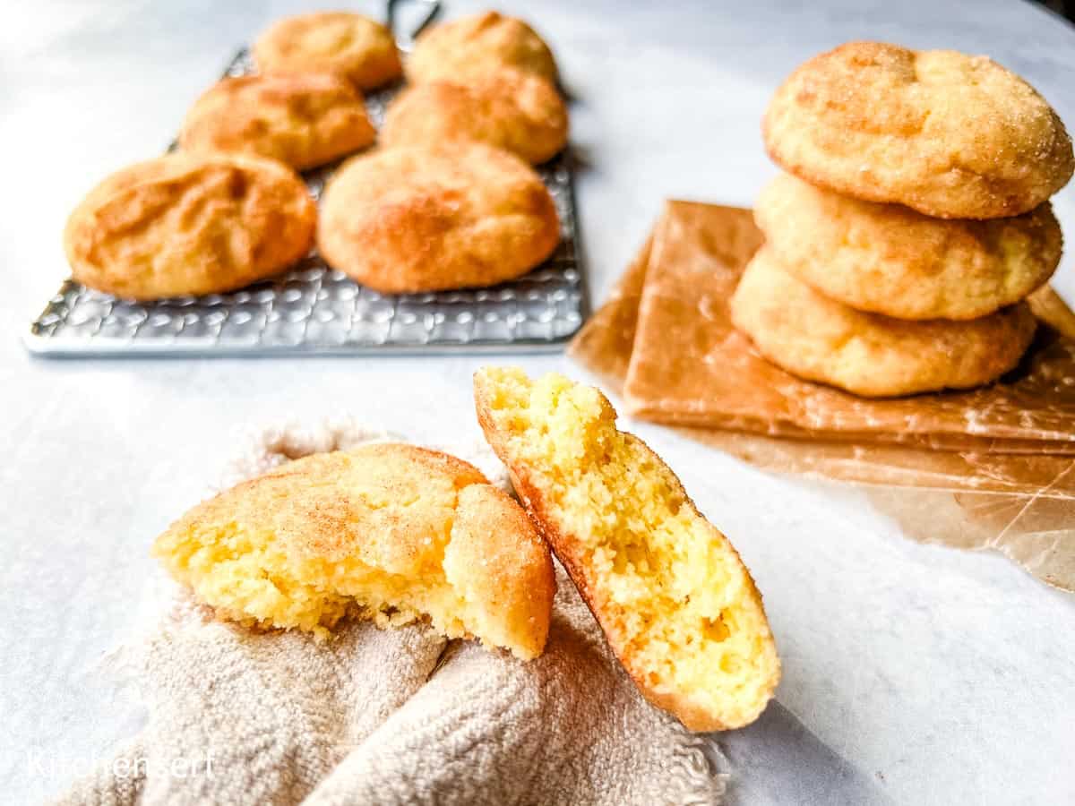 A batch of golden snickerdoodle cookies, some stacked on parchment, others on a cooling rack, with one cookie broken open to show its soft, fluffy interior.