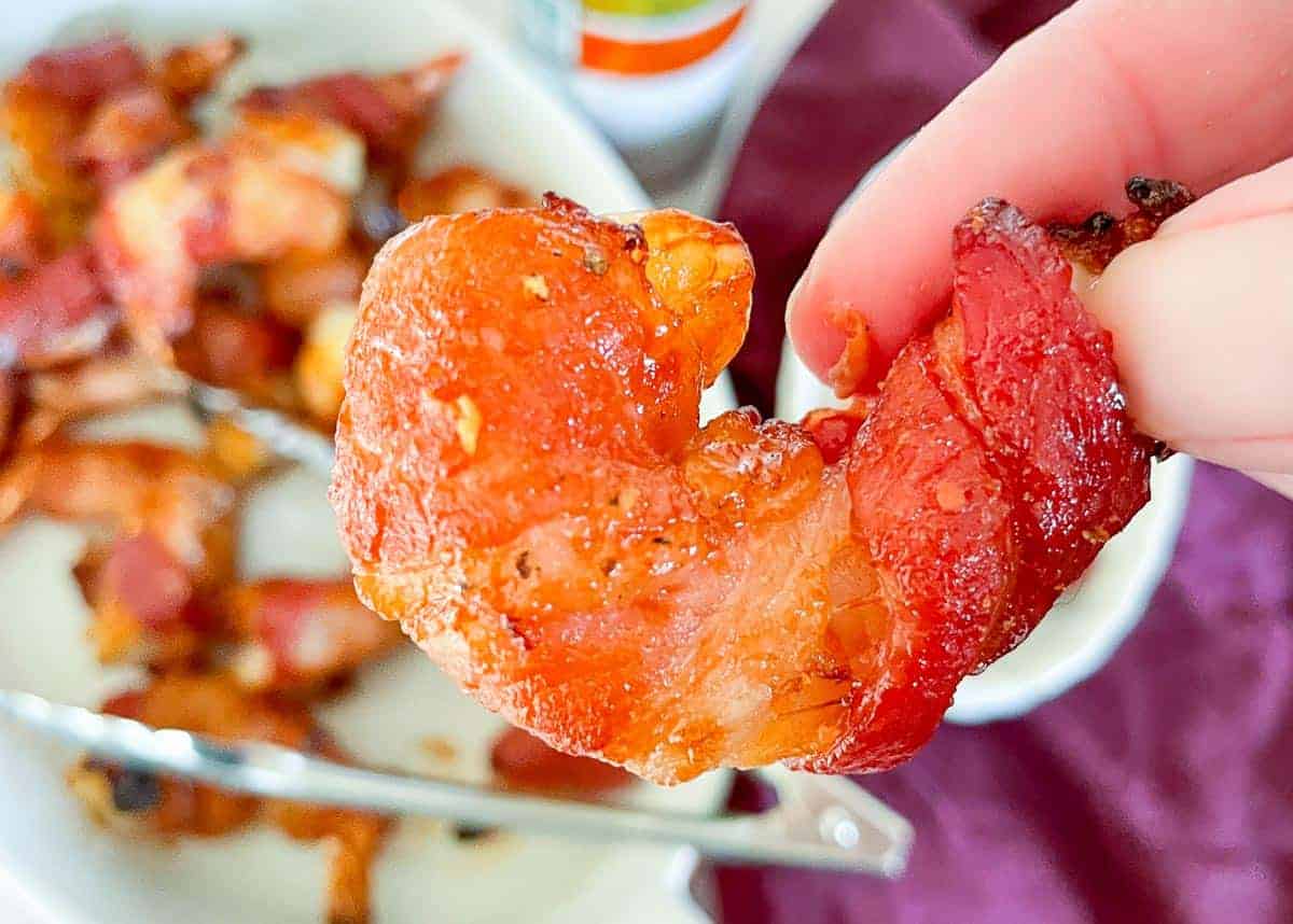 A hand holding a piece of cooked bacon shaped like a heart, with a plate of more bacon pieces in the background.