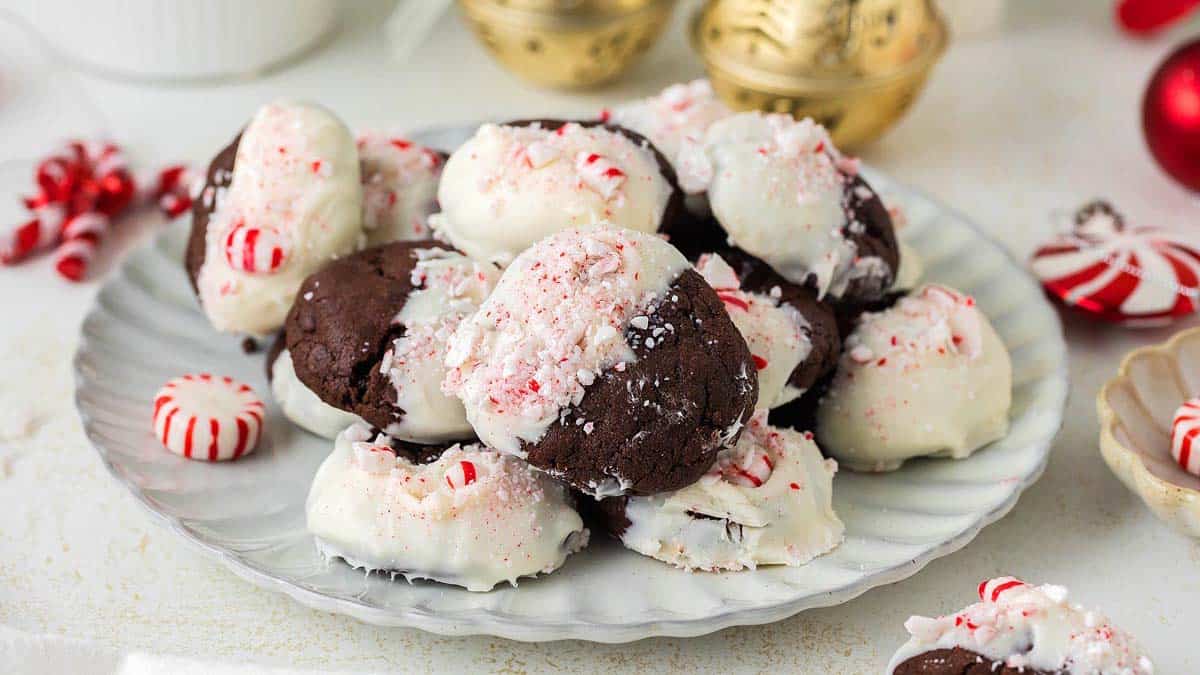 A plate of chocolate cookies half-dipped in white chocolate and sprinkled with crushed peppermint candies, surrounded by festive holiday decorations.