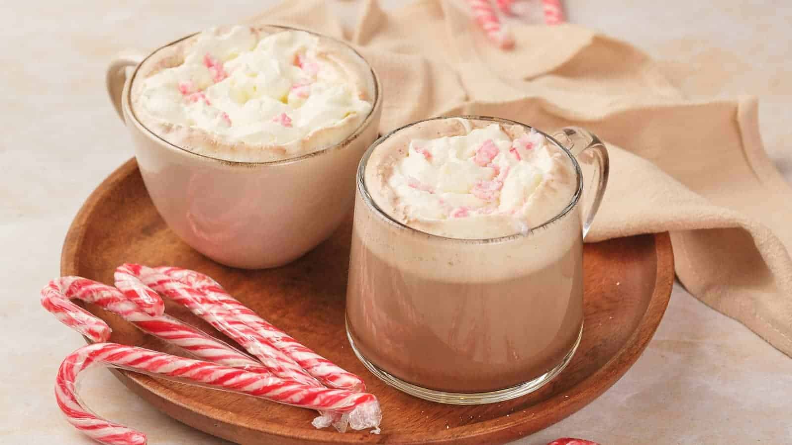 Two mugs of hot chocolate topped with whipped cream and crushed candy canes sit on a wooden plate, surrounded by several peppermint candy canes.
