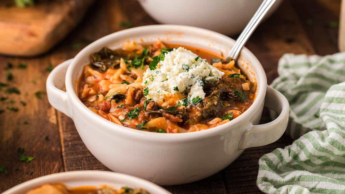 A white bowl of tomato-based soup with rice, vegetables, greens, and a scoop of cheese on top, with a spoon inside, on a wooden table.