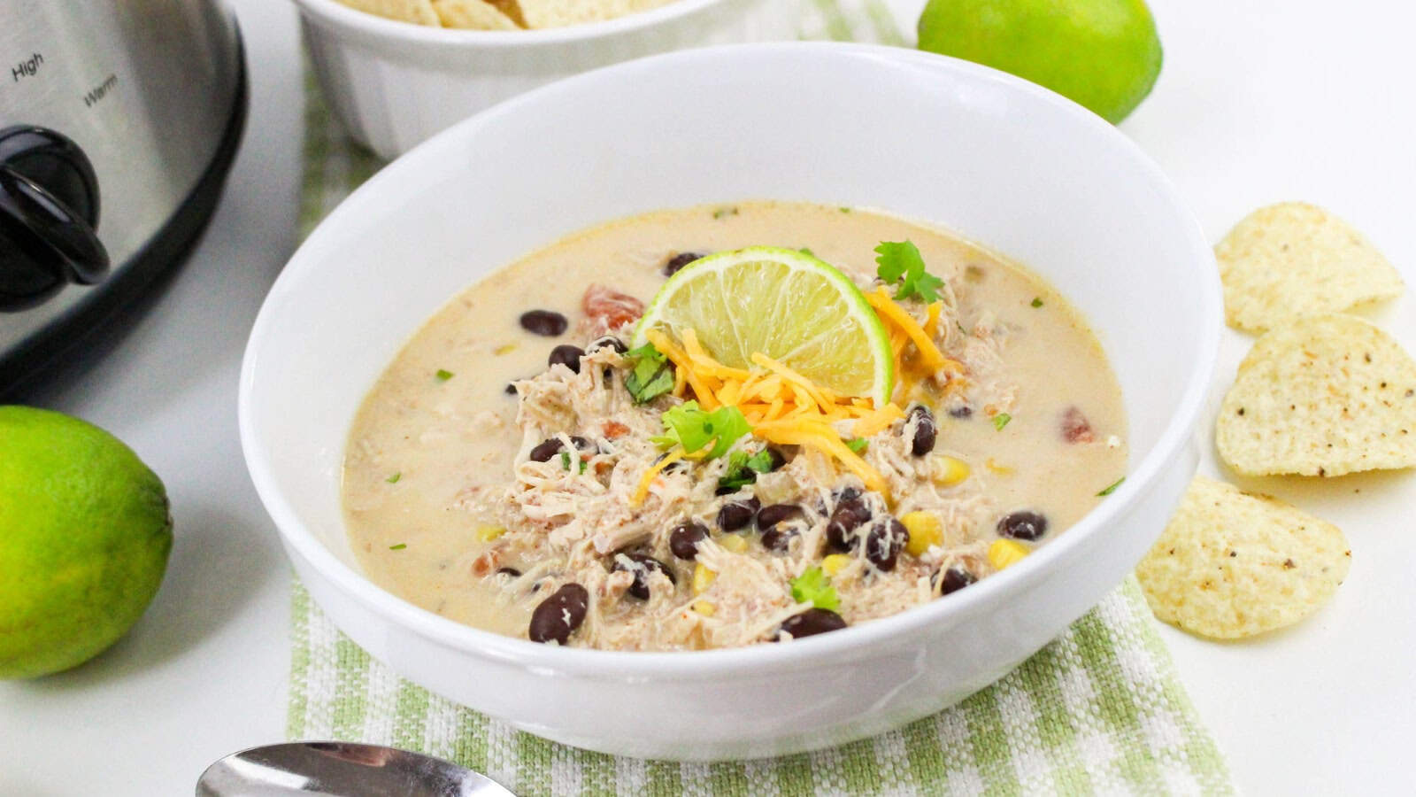A white bowl of creamy chicken chili with black beans, corn, shredded cheese, cilantro, and a lime slice garnish, next to tortilla chips and a lime.