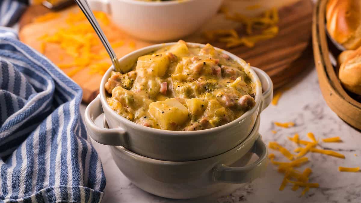 A bowl of creamy potato and broccoli soup with chunks of potato and cheese, served with a spoon, next to shredded cheese and a striped cloth.