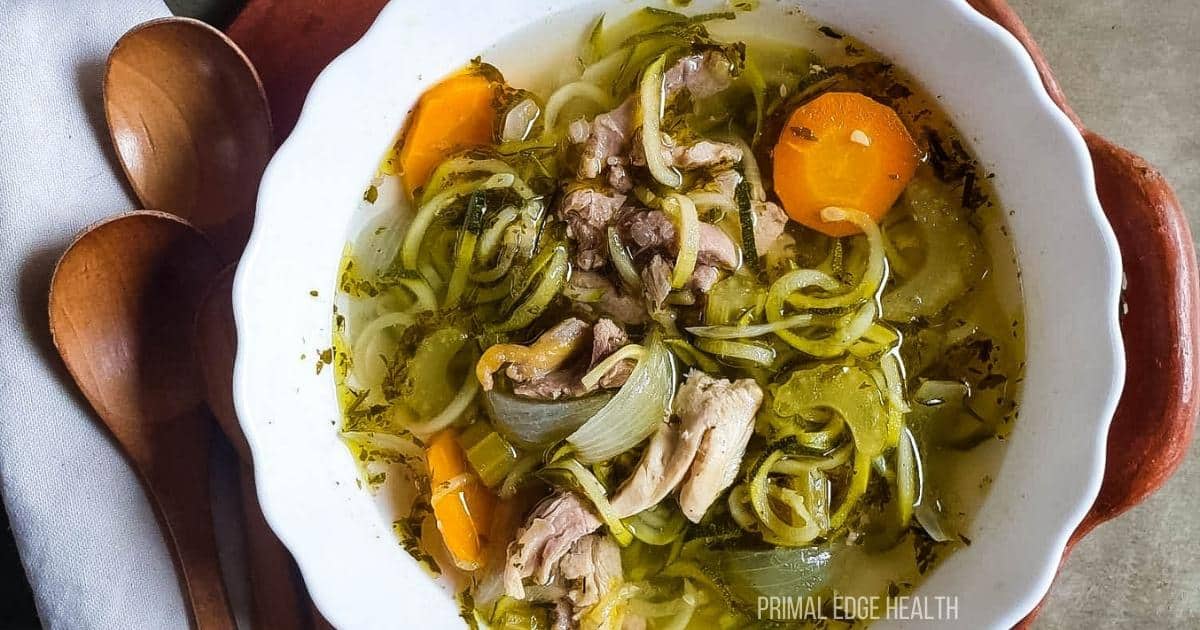 A bowl of chicken soup with shredded chicken, zucchini noodles, carrot slices, onion, and herbs, next to two wooden spoons.