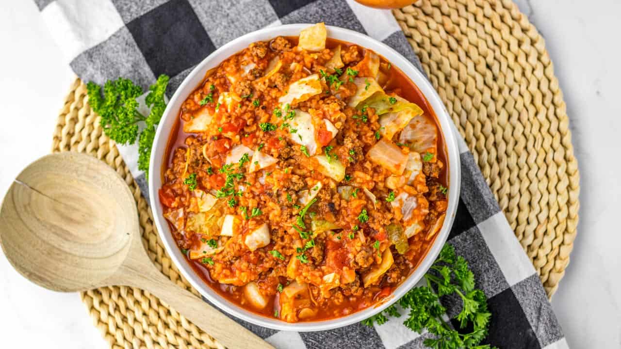 A bowl of cabbage roll casserole with ground meat, tomato sauce, and cabbage pieces, garnished with parsley, sits on a woven placemat with a wooden spoon nearby.