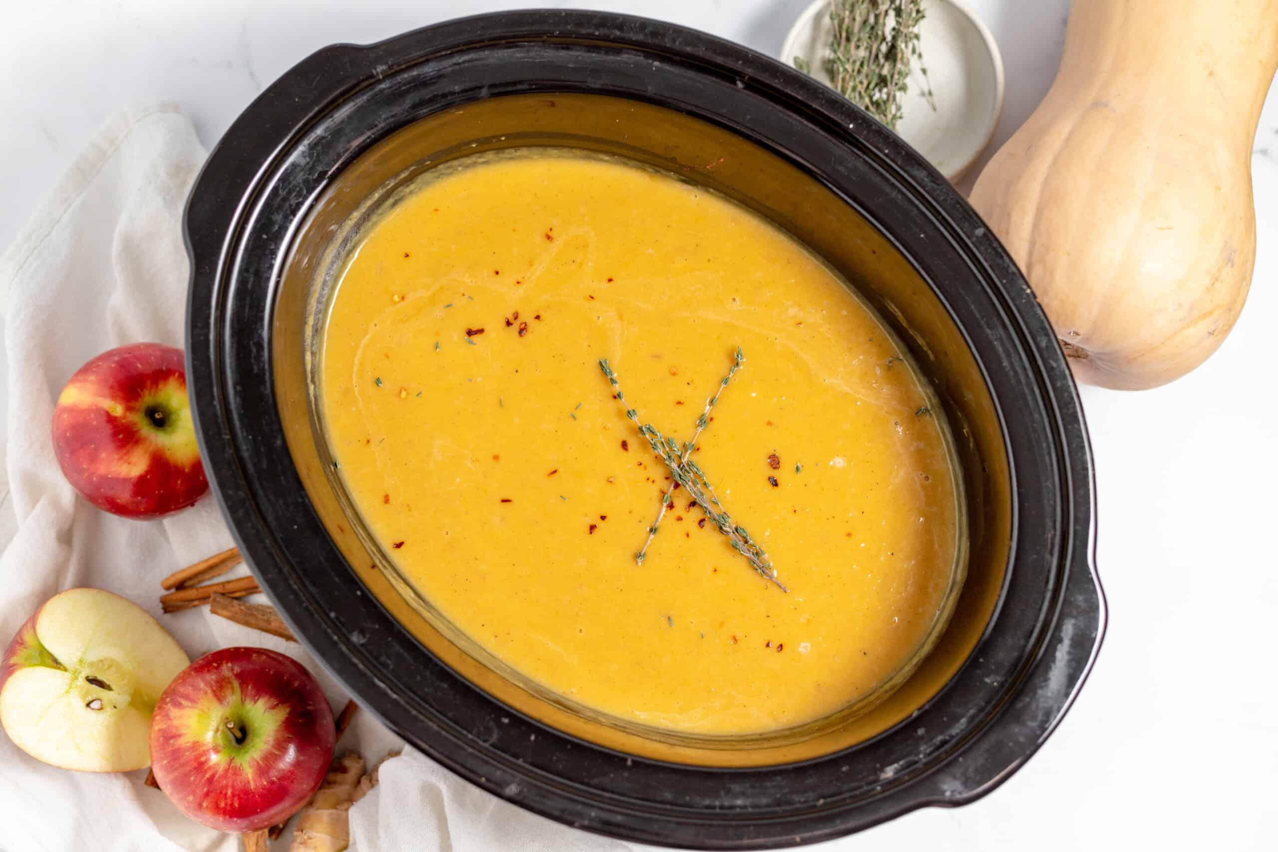 A slow cooker filled with orange-colored soup, garnished with herbs, surrounded by apples, a butternut squash, and cinnamon sticks on a white surface.