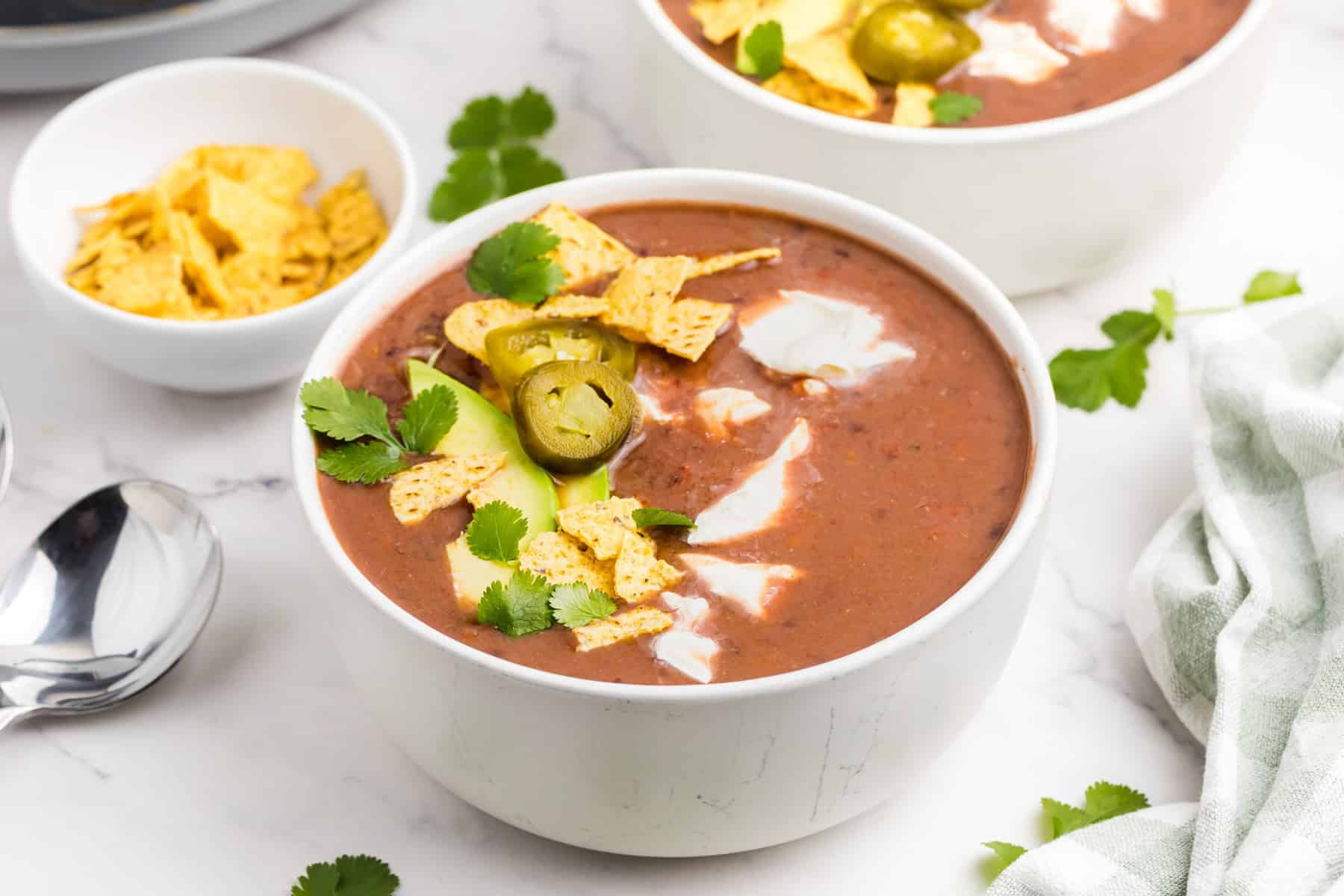 A bowl of black bean soup topped with tortilla chips, sour cream, jalapeño slices, and cilantro, with a small bowl of chips nearby.