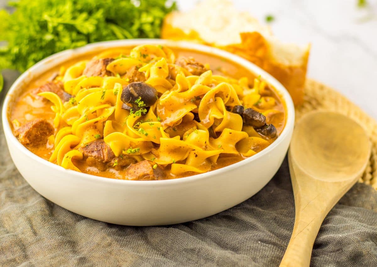 A bowl of noodle stew with pieces of meat and mushrooms, garnished with herbs, sits on a cloth next to a wooden spoon and bread.