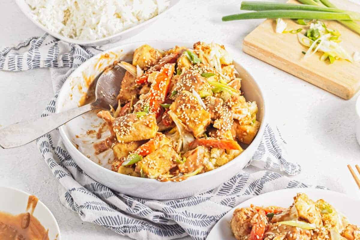 A bowl of stir-fried tofu with vegetables and sesame seeds sits on a patterned cloth, with rice and chopped scallions in the background.