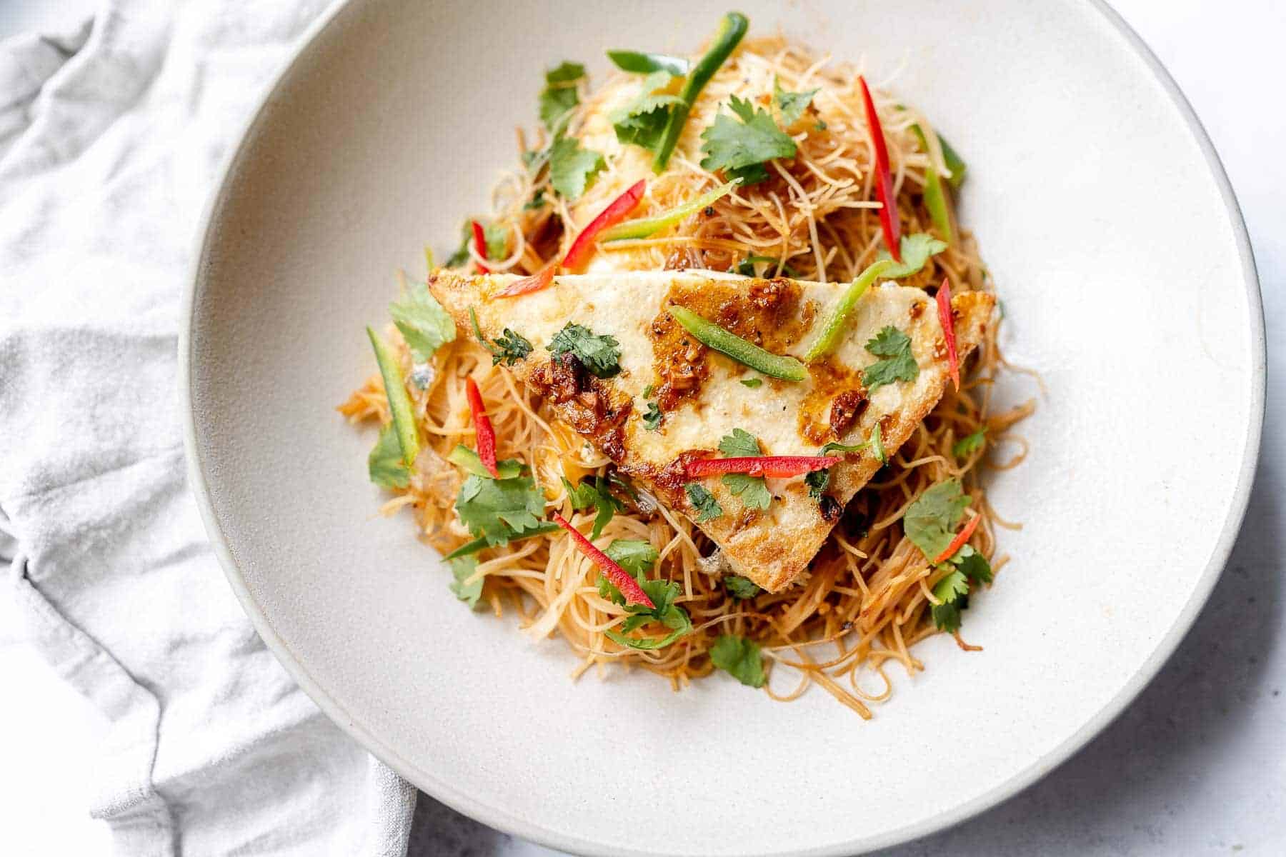 A plate of stir-fried rice noodles topped with tofu triangles, garnished with cilantro and sliced red and green peppers.