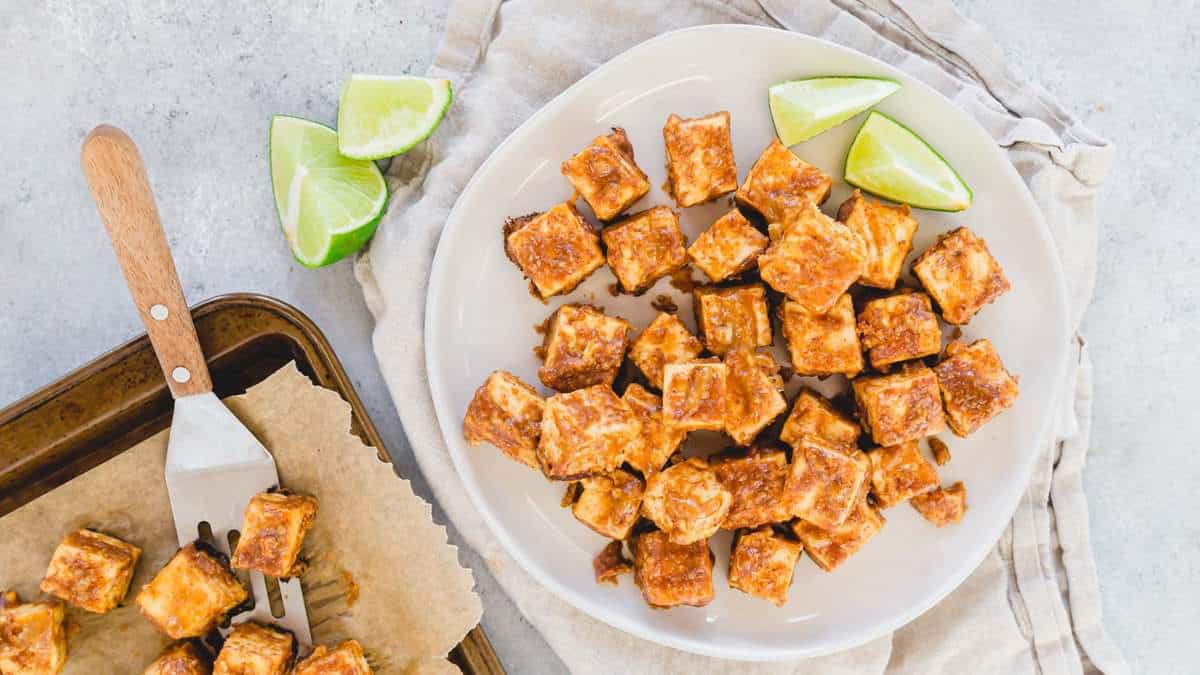 A white plate holds cubed, baked tofu with a golden crust, garnished with lime wedges. Nearby, more tofu cubes rest on a parchment-lined tray with a spatula.