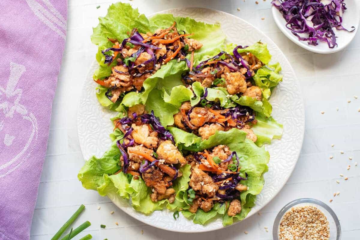 A plate of lettuce wraps filled with cooked chicken, shredded carrots, and purple cabbage, next to a bowl of extra cabbage and a small dish of sesame seeds.