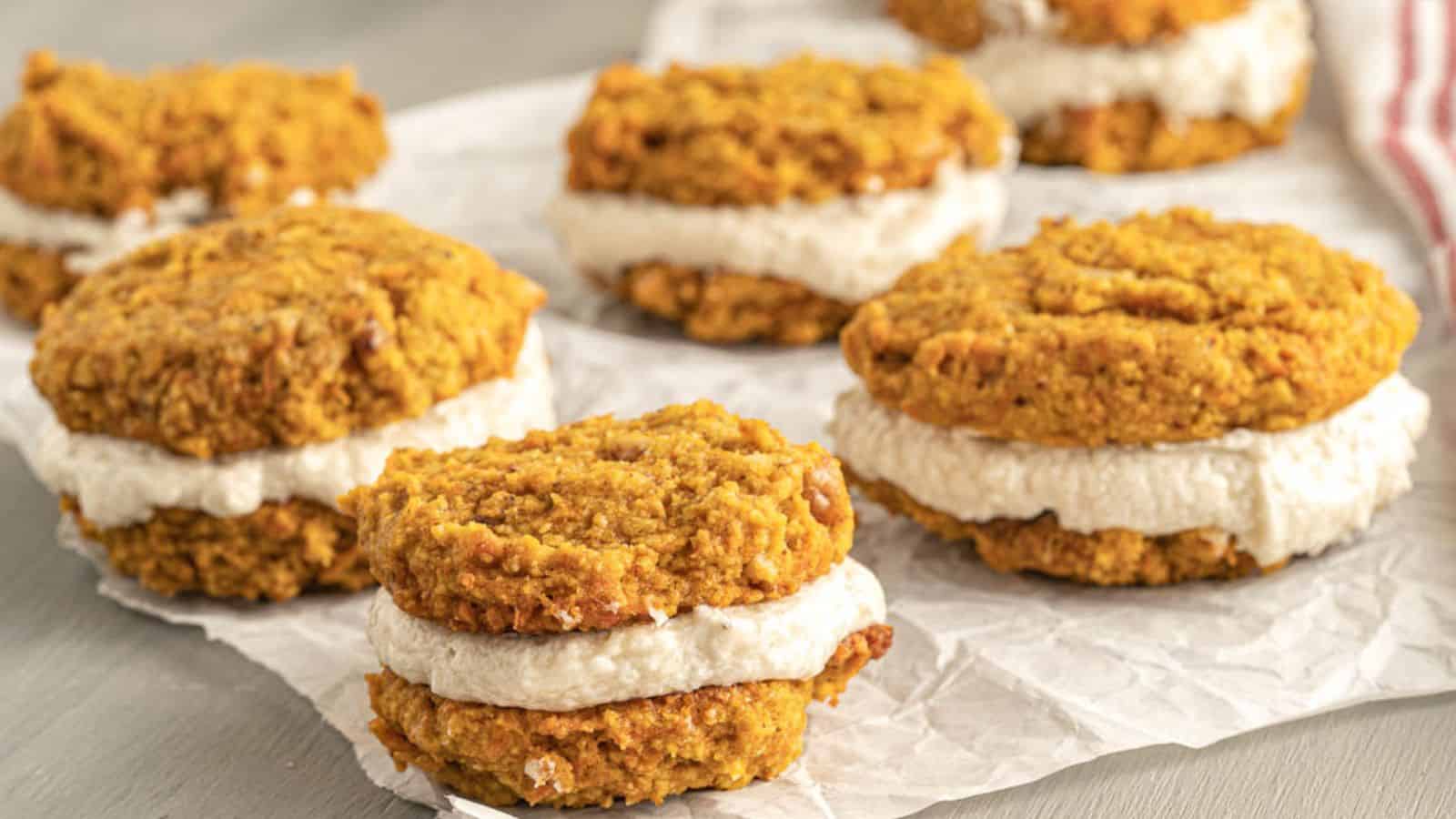 Pumpkin cookie sandwiches with a creamy white filling, arranged on crumpled parchment paper on a light-colored surface.