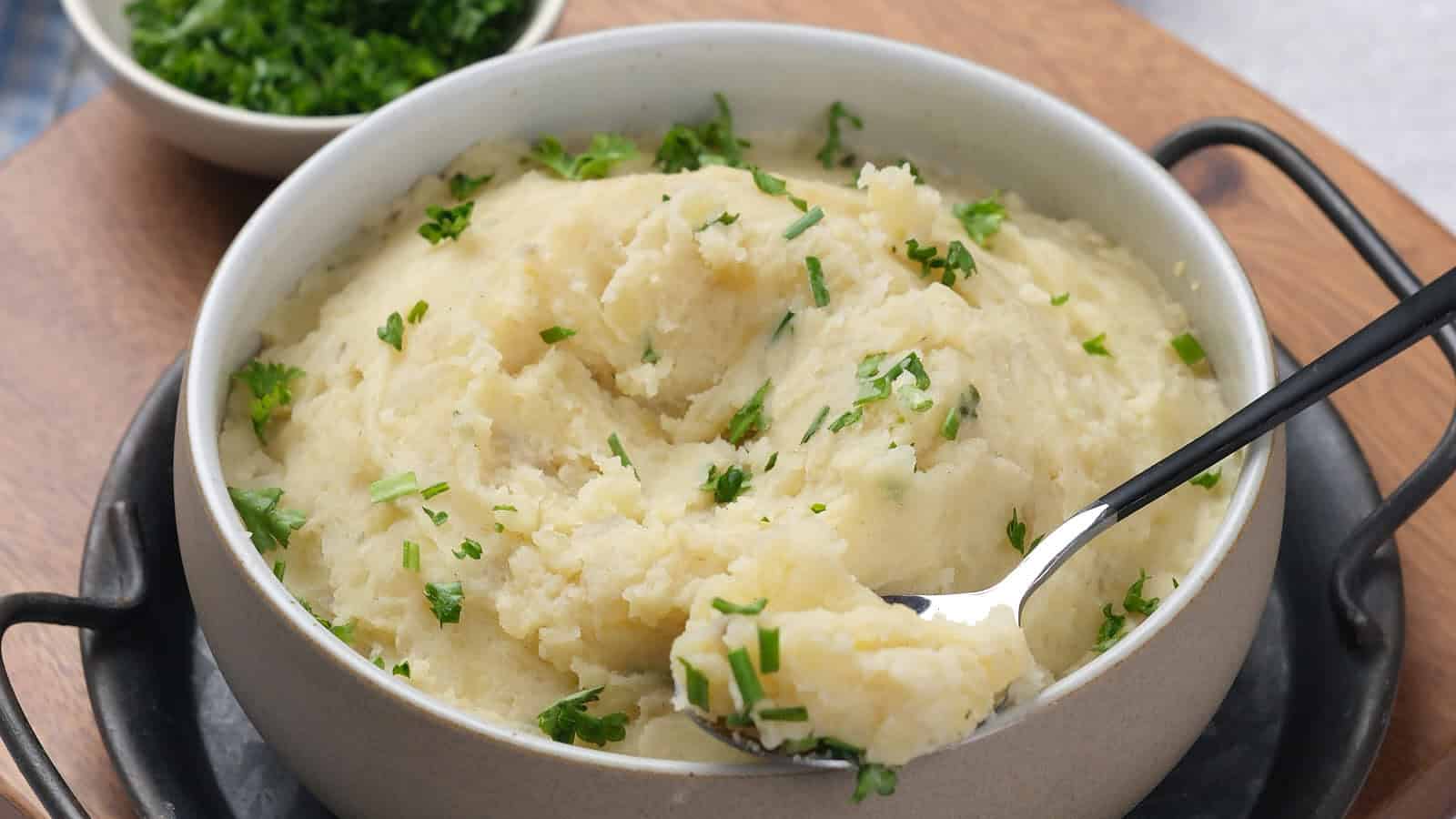 A bowl of mashed potatoes garnished with chopped herbs sits on a tray, with a spoon scooping some out and a small bowl of herbs in the background.