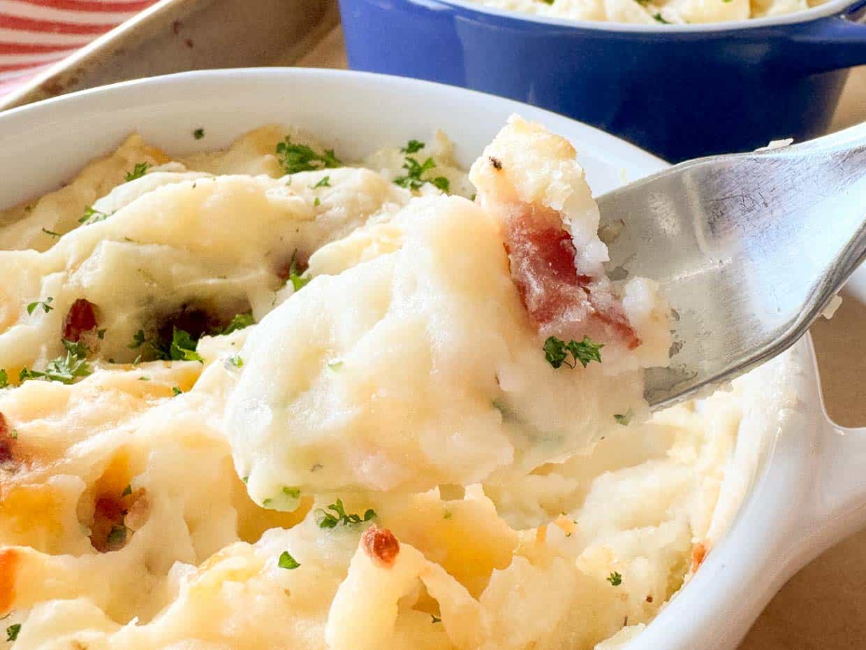 A fork lifts a portion of mashed potatoes with herbs and bacon from a white baking dish, with another blue dish in the background.