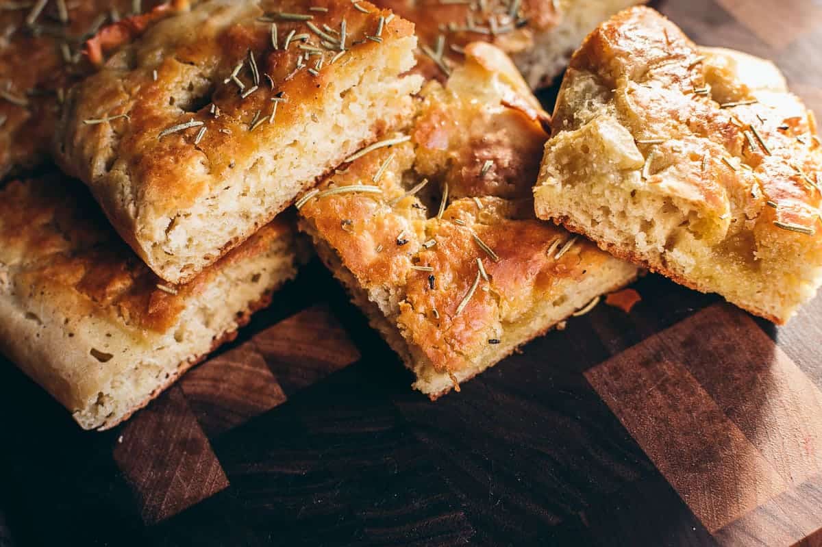Slices of golden-brown focaccia bread topped with herbs are arranged on a dark wooden surface.