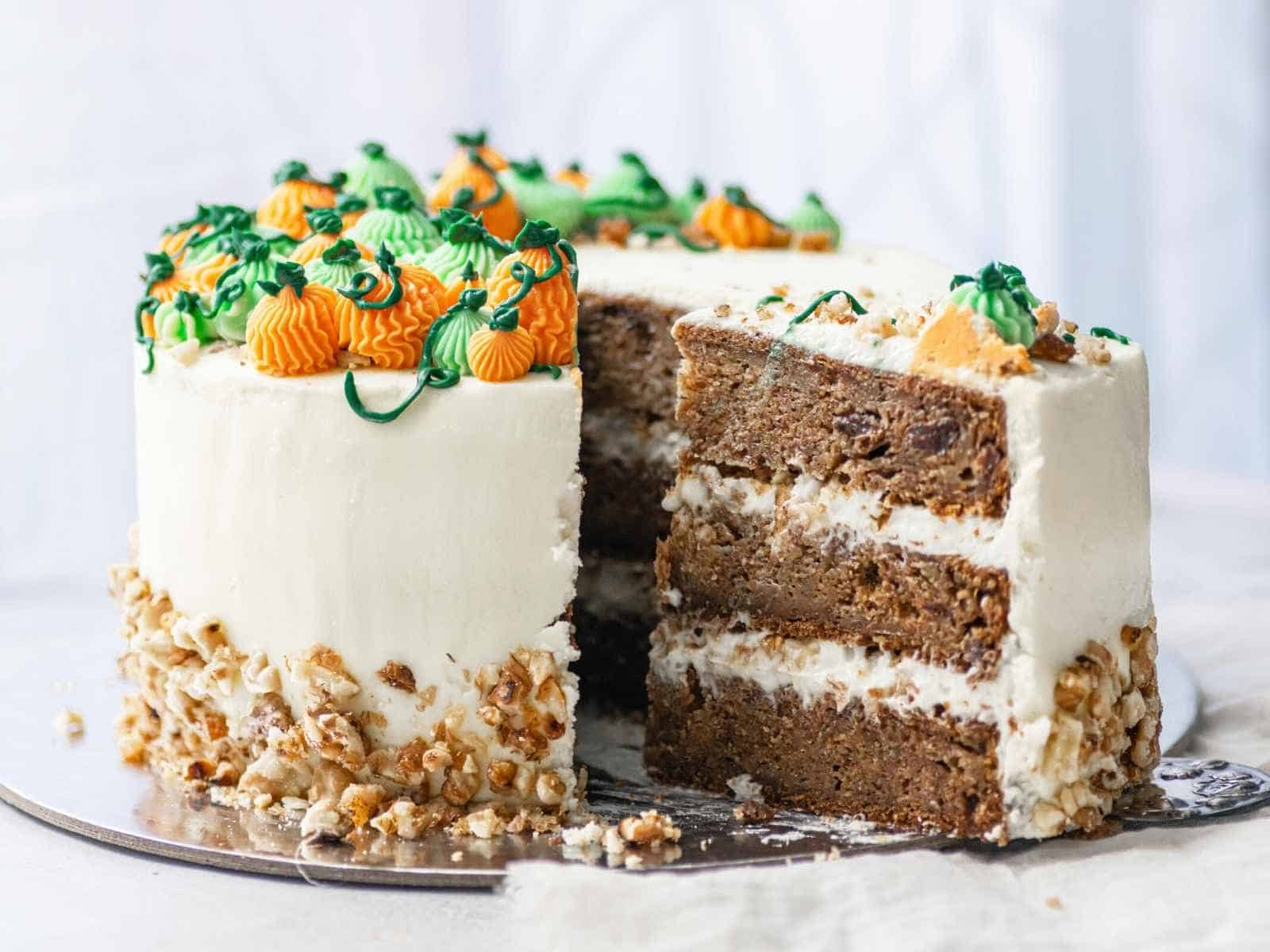 A three-layer carrot cake with white frosting, nut pieces on the sides, and orange and green decorative icing on top; one slice is partially removed.