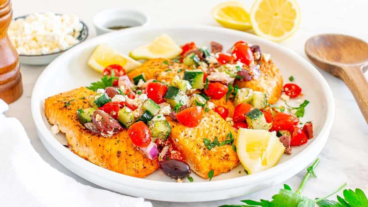 Grilled salmon fillets topped with a Mediterranean salsa of tomatoes, cucumbers, olives, and feta, garnished with lemon wedges on a white plate.