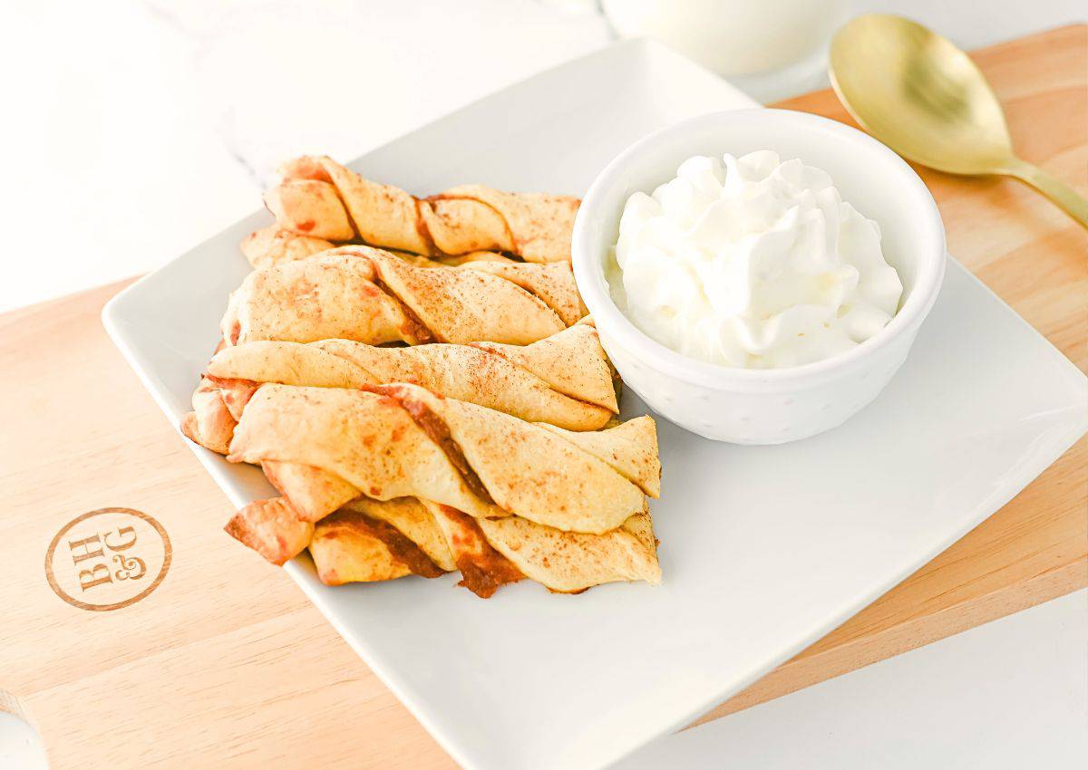 A white plate with cinnamon twists next to a bowl of whipped cream, set on a wooden board with a gold spoon beside it.