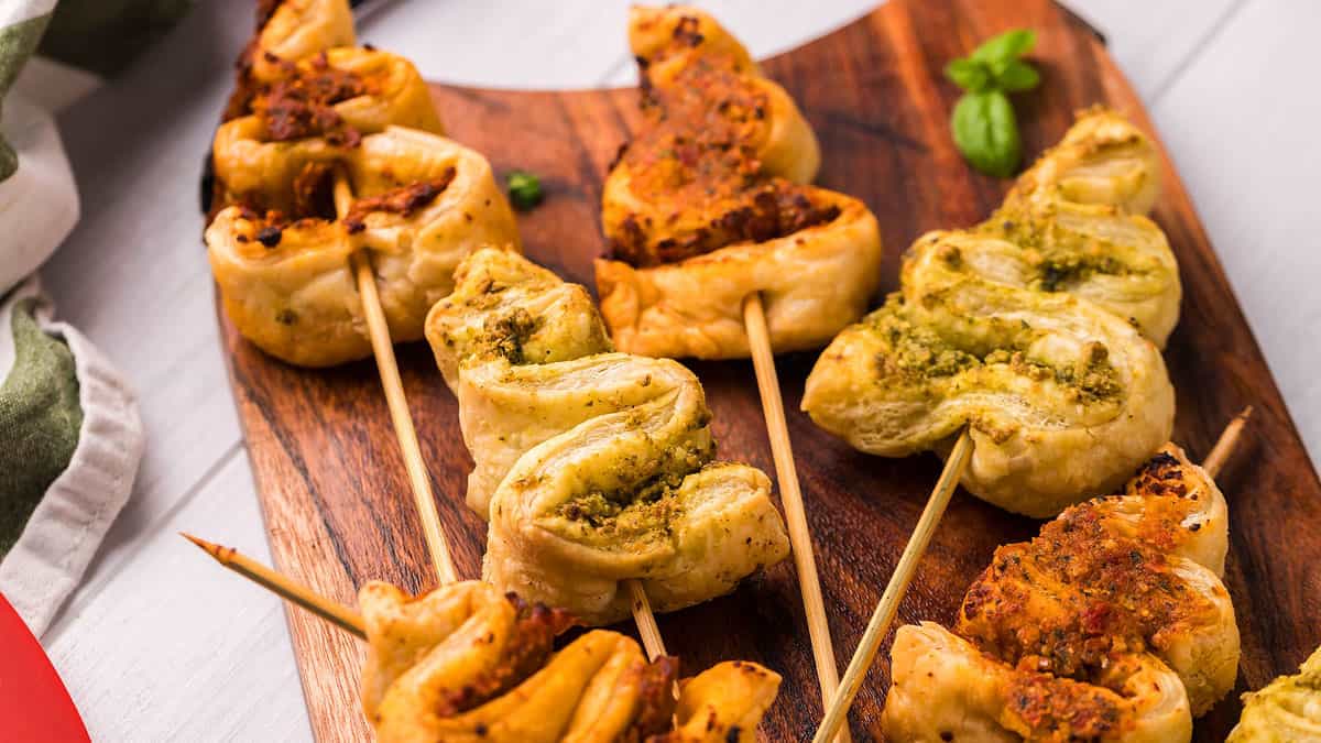 Puff pastry twists with green and red pesto fillings are skewered on sticks and arranged on a wooden serving board.