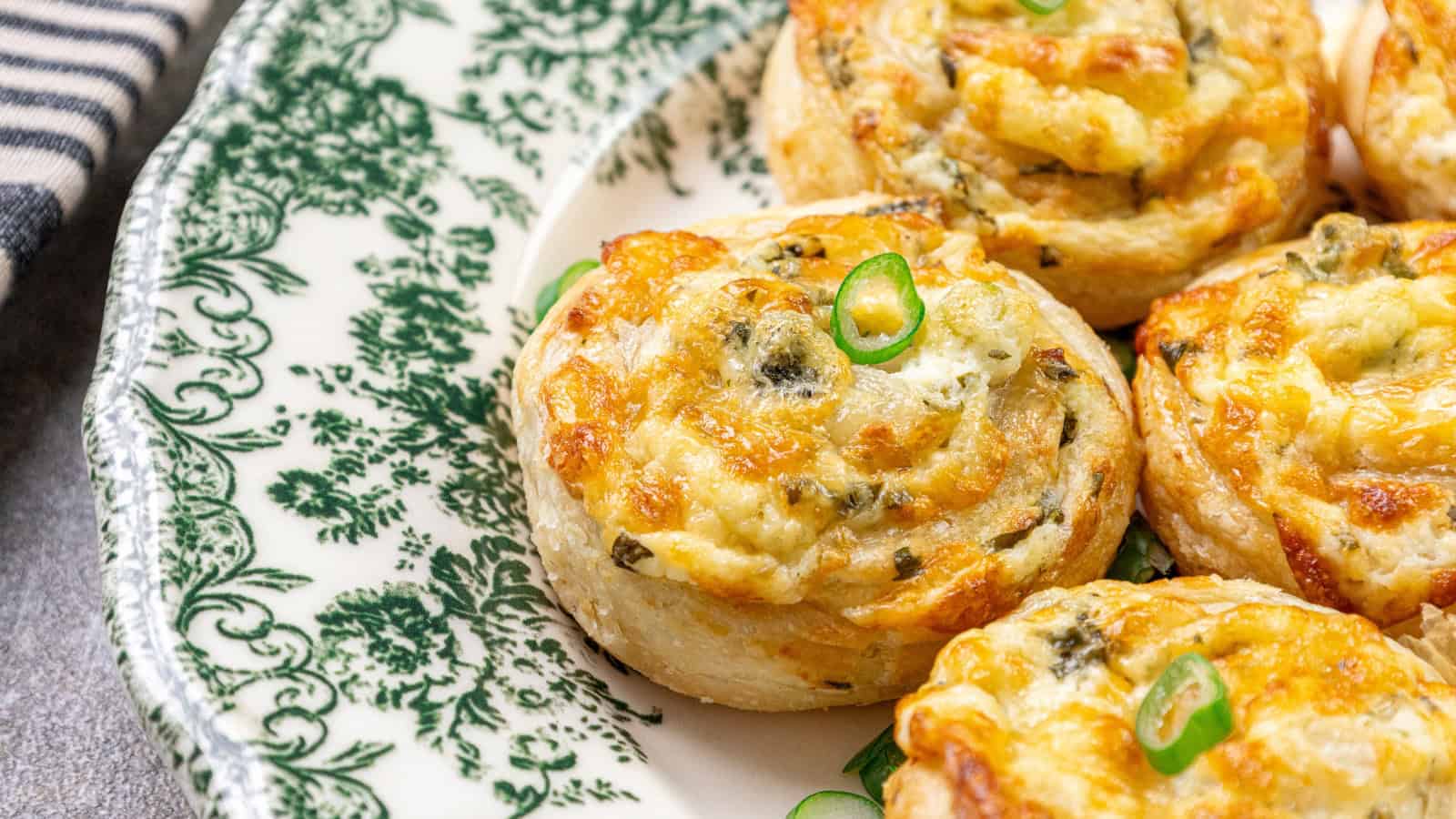 A close-up of savory pastry pinwheels with melted cheese and herbs, garnished with sliced green onions, served on a decorative green and white plate.