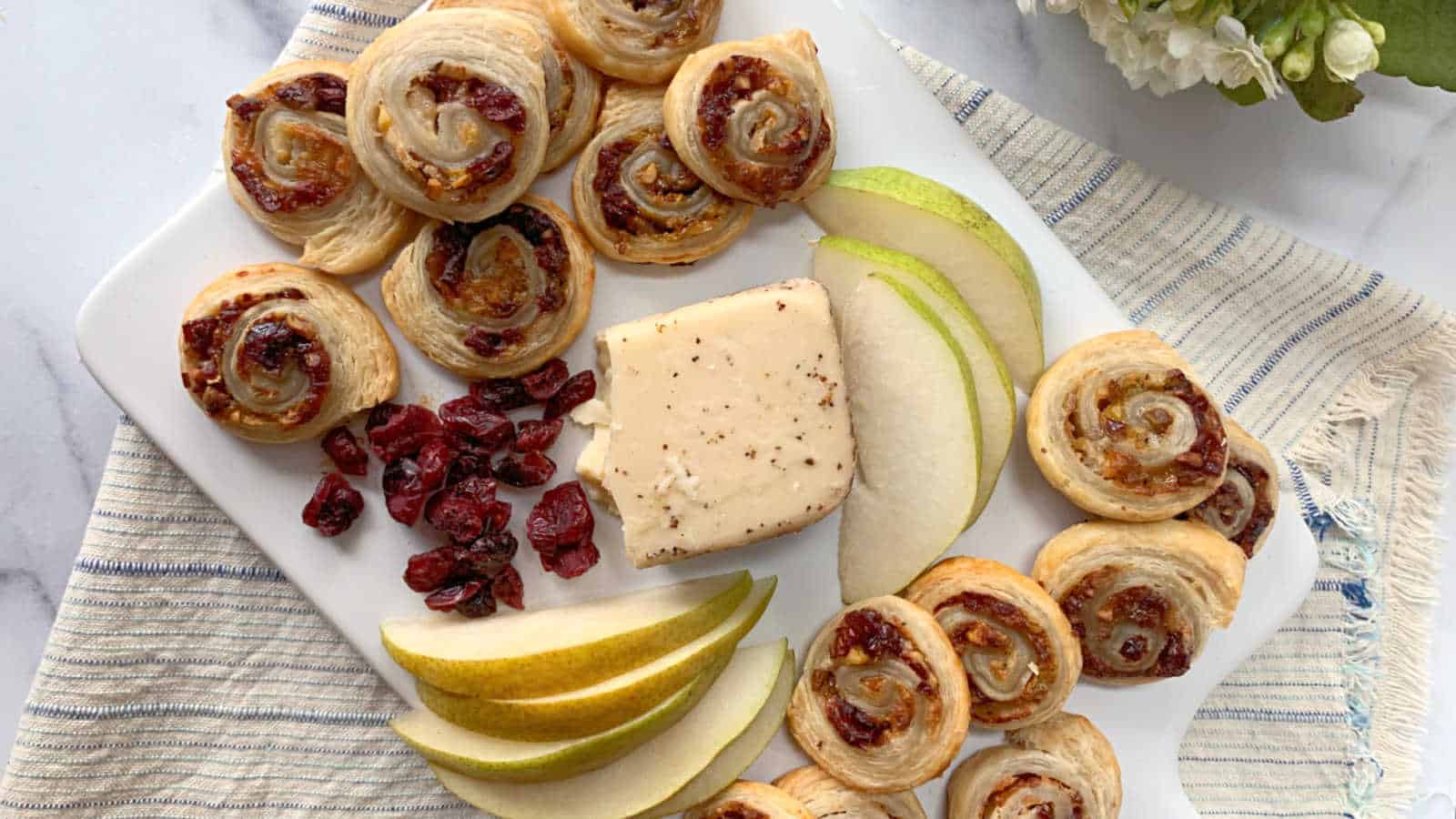 A white platter with puff pastry pinwheels, dried cranberries, a block of cheese, and sliced pears, arranged on a striped cloth.
