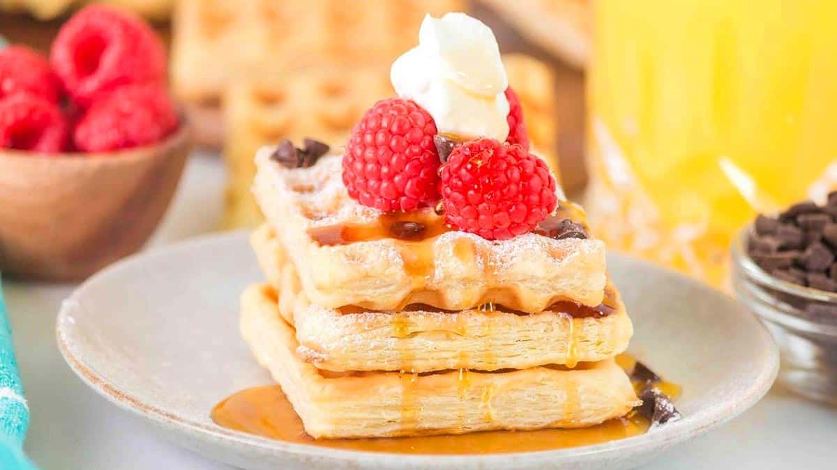 A stack of waffles topped with raspberries, whipped cream, chocolate chips, and syrup sits on a plate; raspberries and chocolate chips are in bowls nearby.
