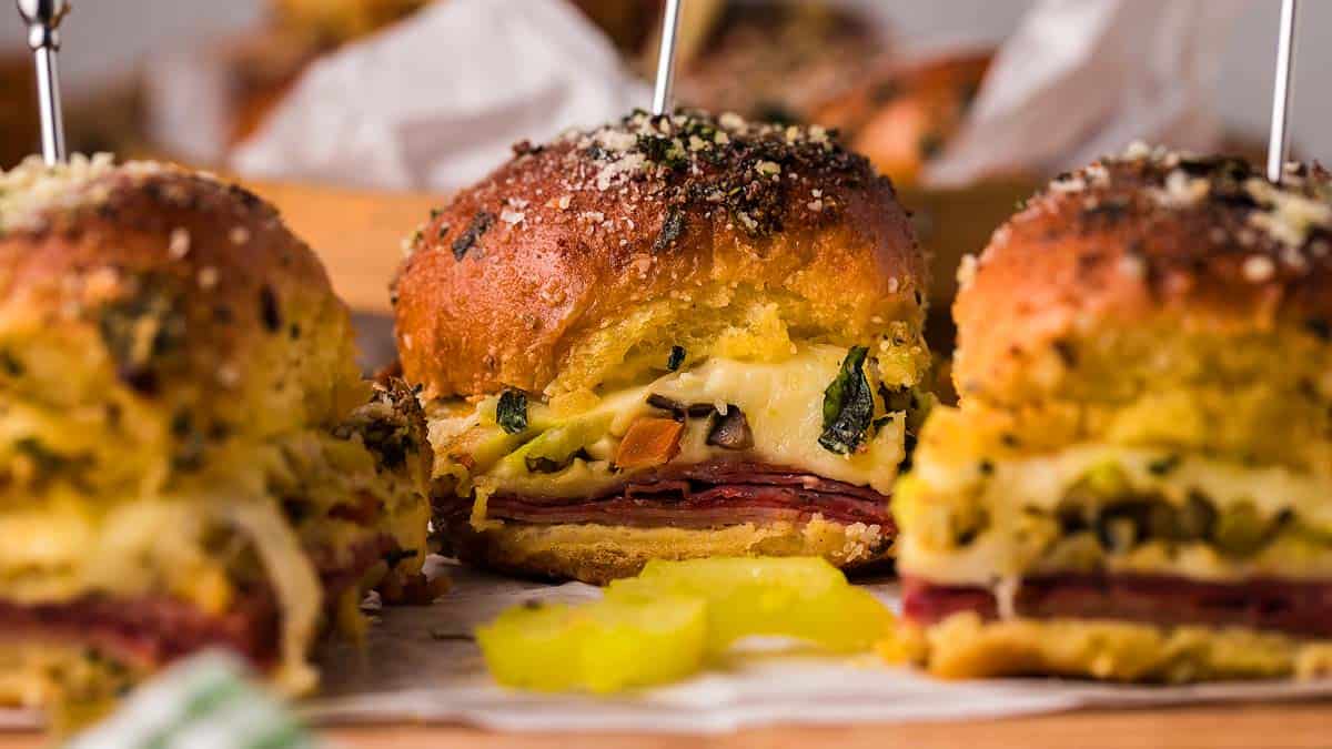 Close-up of baked sandwich sliders filled with layers of meat, cheese, vegetables, and herbs, topped with grated cheese and served with pickle slices.